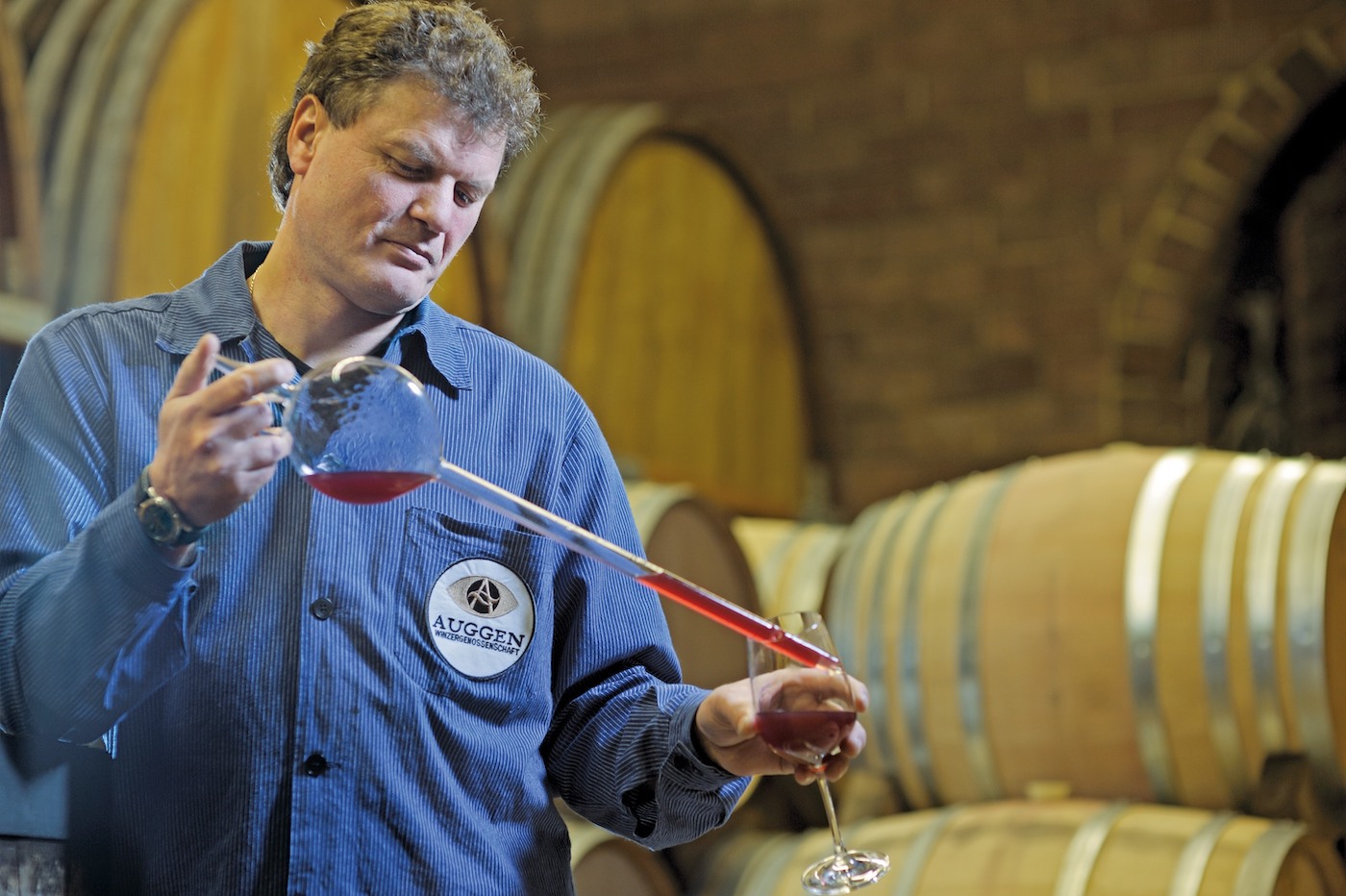 Kellermeister Andreas Philipp zieht eine Probe aus einem Rotweinglas, im Hintergrund aufeinandergestapelte Weinfässer in einem Keller.