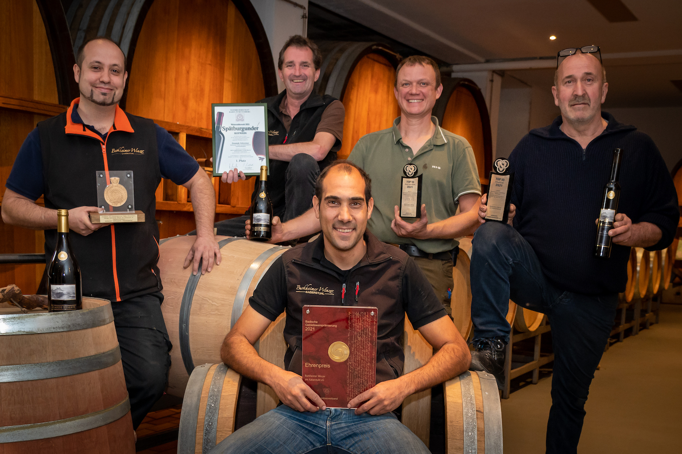 Fünf Männer stehen mit diversen Medaillen uns Auszeichnungen in einem Weinkeller vor großen Weinfässern und lachen stolz in die Kamera.