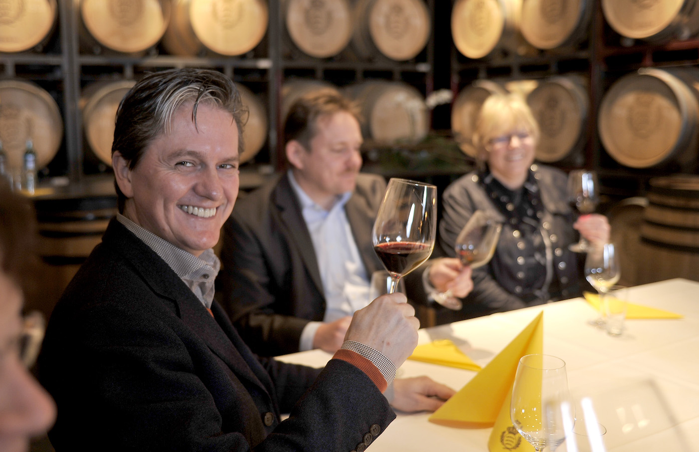 Drei mittelalte Personen sitzen um einen fein mit Servietten eingedeckten Tisch und verkosten Rotwein, im Hintergrund zahlreiche Weinfässer aus Holz.