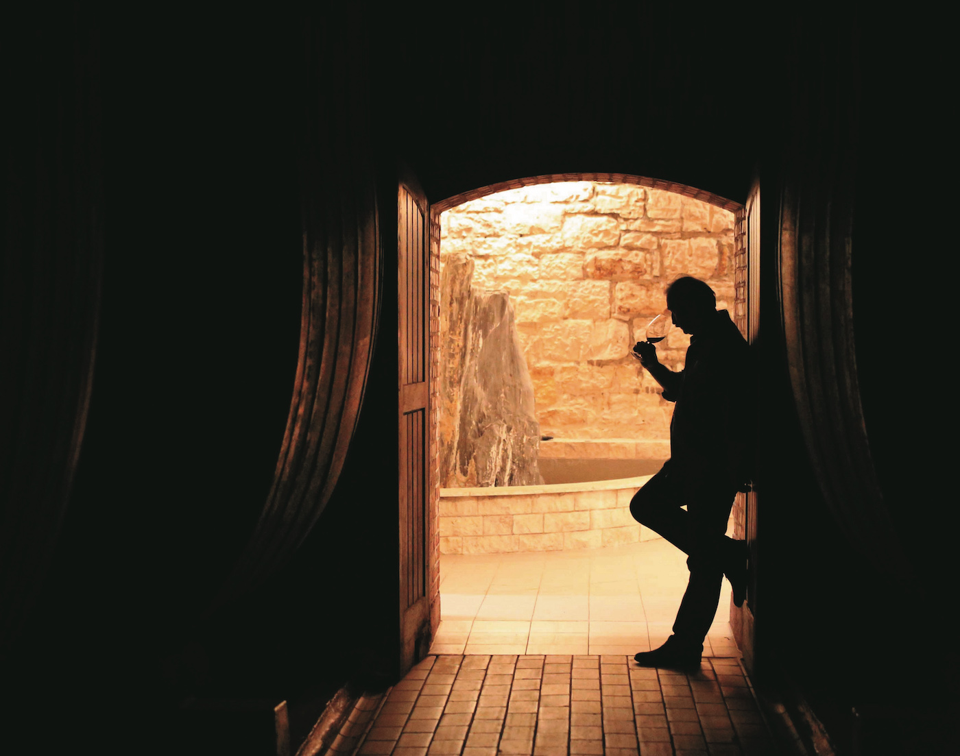 Blick aus einem dunklen Raum in einen hellen Weinkeller, im Gegenlicht steht ein Mann im Tor, der in sein Rotweinglas blickt.